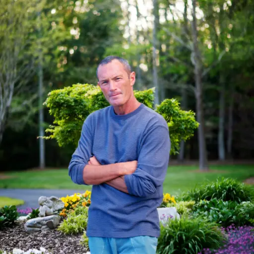 man standing with arms crossed in a landscaped garden, surrounded by greenery and colorful flowers.